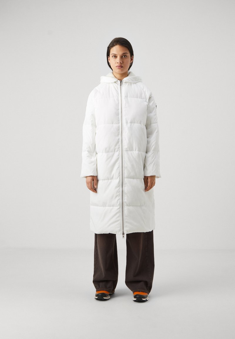 Doudoune blanche avec capuche, dotée d'une fermeture éclair intégrale et de sections matelassées horizontales. Portée par-dessus un pantalon large sombre et des baskets.