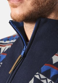 Marinefarbener Zip-Pullover mit geripptem Muster, ausgestattet mit einem blauen Reißverschluss und einem geometrischen Muster in Blau, Rot und Weiß auf der Schulter.
