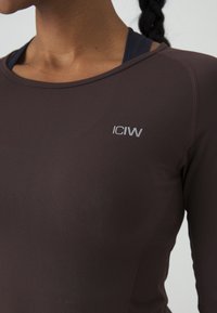 Brūns garo piedurkņu sporta tops, izgatavots no gludas materiāla, ar apaļo dekoltē un mazu sudraba logotipu uz krūšu daļas.