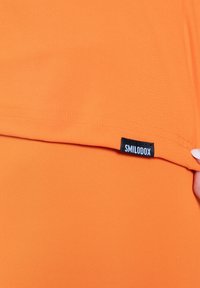 Camiseta deportiva naranja hecha de tela suave y elástica con una etiqueta negra "SMILODOX" en la costura. Presenta un diseño sencillo sin estampados.