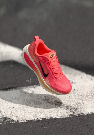 Jaskrawy koralowy but do biegania z czarnym logo swoosh, białą podeszwą i etykietą „RUN”, zawieszony nad asfaltem z białymi znakami drogowymi.