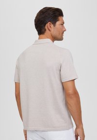 Camisa polo de manga corta en beige claro con cuello, que presenta una tela texturada y aberturas laterales en el dobladillo para mayor facilidad de movimiento.