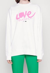 Sudadera blanca de corte oversized con el texto rosa "AMA A TU VICHO" y un detalle de corazón negro, combinada con pantalones verdes. Material suave y ajuste relajado.