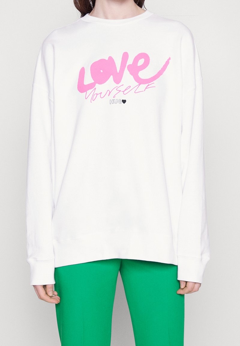 Sudadera blanca de corte oversized con el texto rosa "AMA A TU VICHO" y un detalle de corazón negro, combinada con pantalones verdes. Material suave y ajuste relajado.