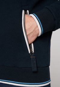 Felpa con zip blu navy con tasca interna bianca, polsini a costine con strisce blu e bianche, e una cintura testurizzata.