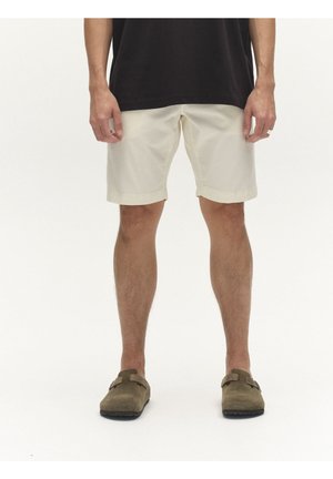 Bărbat purtând pantaloni scurți bej, pantofi slip-on verde măsliniu și o cămașă neagră, stând pe un fundal alb simplu.