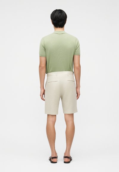 Άνδρας που φοράει ανοιχτό πράσινο πόλο, μπεζ σορτς και μαύρα σανδάλια, στέκεται γυρισμένος με την πλάτη προς τα μπρος σε απλό φόντο.