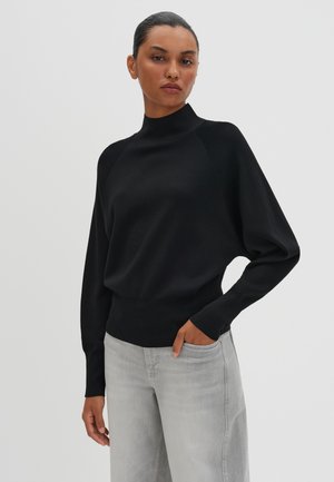 Schwarzer gestrickter Pullover mit hohem Kragen, Raglanärmeln und schmalem Bund, kombiniert mit hellgrauen, weit geschnittenen Jeans.