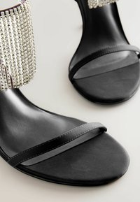 Mustad kontsad mustadest nahast, millel on sile nahk, laia rihmaga ja kaunistatud ankle cuff, mis on kaetud langevate rhinestones'idega.
