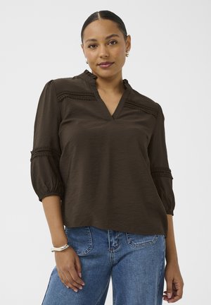 Femme portant un chemisier marron foncé avec des détails en dentelle et des manches trois-quarts, associé à un jean bleu, posant devant un fond uni.