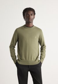 Maglione a trama verde oliva con scollo a girocollo, polsini e orlo a costine, dotato di una piccola etichetta logo sul petto sinistro. Abbinato a pantaloni neri.