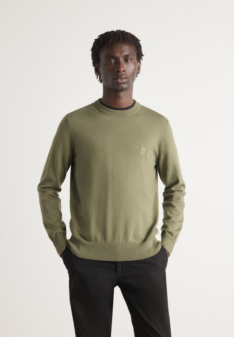 Maglione a trama verde oliva con scollo a girocollo, polsini e orlo a costine, dotato di una piccola etichetta logo sul petto sinistro. Abbinato a pantaloni neri.