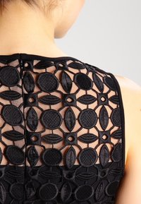 Svart ärmlös klänning med en texturerad broderad rygg med cirkulära och ovala mönster på ett genomskinligt meshmaterial. Dragkedjedetalj ingår.