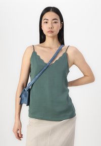 Green sleeveless top with scalloped neckline, paired with a textured beige skirt. Blue strap crossbody bag rests on one shoulder.
