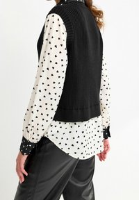 Canottiera nera a costine sopra una camicia bianca con motivi a cuori neri, abbinata a pantaloni in pelle nera. Presenta maniche lunghe con dettagli ai polsini.