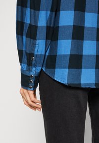Blue and black plaid flannel shirt with long sleeves, buttoned cuffs, and a curved hem, paired with dark denim pants.
