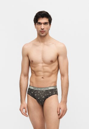 Utrzymany w dobrej formie mężczyzna z krótkimi ciemnymi włosami, ubrany w ciemne slipy Emporio Armani z wzorzystymi nadrukami logo, stojący na jednolitym jasnym tle.