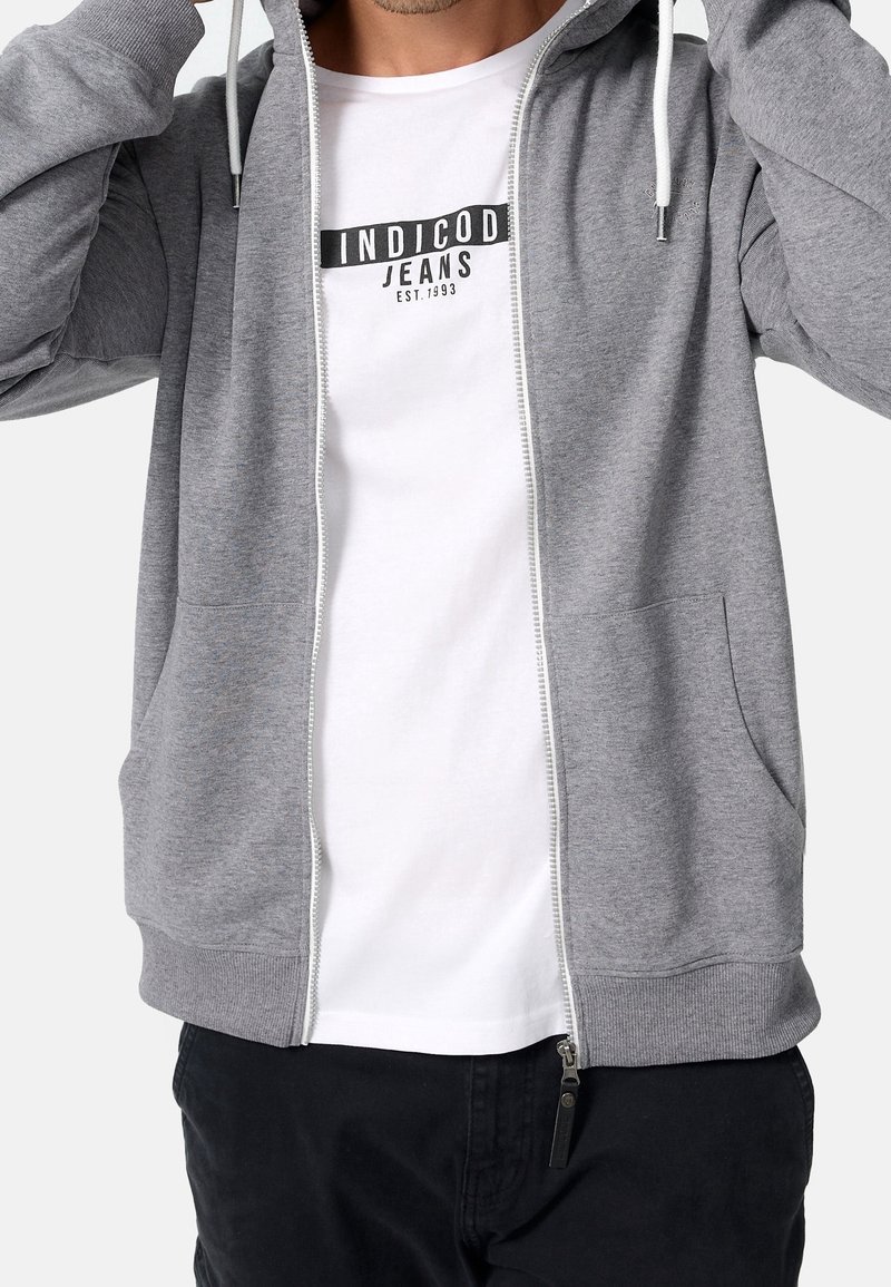 Sweat à capuche gris avec fermeture éclair, texture jersey, cordons, deux poches avant, et un t-shirt blanc avec un logo de marque en dessous.