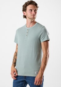 Ljusgrön kortärmad henleytröja med tre träknappar, strukturerat tyg och en liten logotypmärke vid fållen. Matchad med blå jeans.