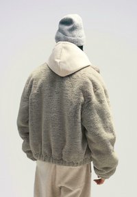 Jachetă bomber bej pufoasă cu glugă crem, talie elastică și manșete acoperite cu dantelă. Purtată cu o pereche de pantalon bej asortată și o căciulă gri.