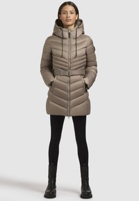 Beige gewatteerde jas met een capuchon, chevron-quilten, een bijpassende riem op de taille, een front-rits en zijzakken, gedragen over zwarte leggins en laarzen.