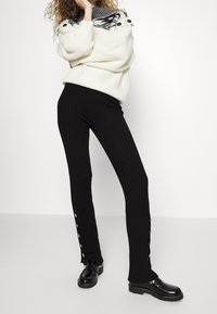 Vit ribbad stickad tröja med en svart mönstrad krage; kombinerad med svarta ribbade byxor med sidoknappar; glänsande svarta skor.