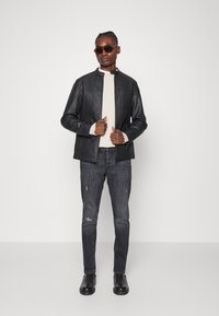 Casaco de pele preto com colarinho levantado, usado sobre um suéter bege. Jeans pretos desgastados e botas pretas completam o olhar casual.