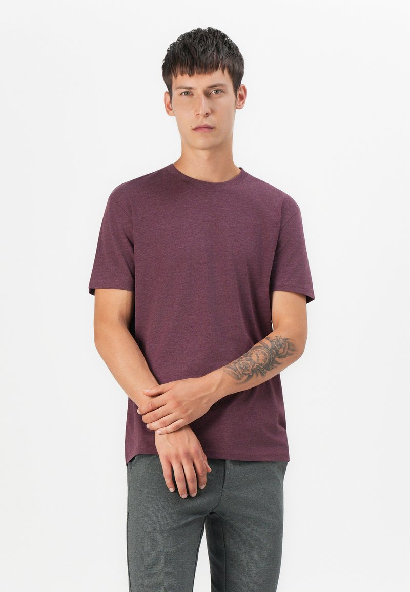 Bordeauxfarbendes kurzärmliges T-Shirt aus einem weichen, strukturierten Stoff, Rundhalsausschnitt, lässiger Schnitt, kombiniert mit grauen Hosen und einem Tattoo am linken Arm.