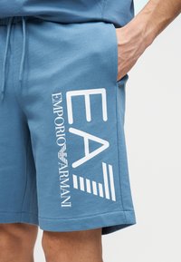 Blå bomullsshorts med avslappnad passform, med vitt "EA"-logotyp och text. Mjuk textur, sidfickor och elastisk midja.