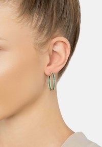 Arany karika fülbevaló zöld és feh ér drágakövekkel, egy nő bal fülén, rövid hajjal és világos bőrrel, fehér háttér előtt.