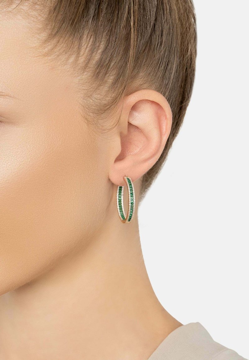Arany karika fülbevaló zöld és fehér drágakövekkel, egy nő bal fülén, rövid hajjal és világos bőrrel, fehér háttér előtt.