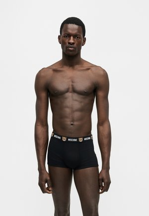 Hombre de piel oscura en forma de pie, vistiendo calzoncillos boxer negros Moschino con logotipos de ositos de peluche y el nombre de la marca en la cintura sobre un fondo liso.