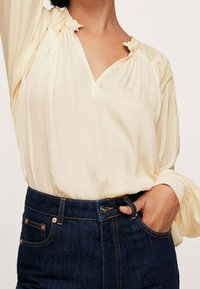 Blusa gialla chiaro con scollo a V e dettagli di colletto arricciato, abbinata a jeans in denim scuro a vita alta con accenti in rame.