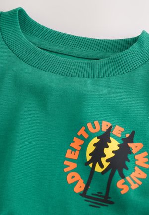Sweat-shirt en coton vert avec un col côtelé. Présente un motif graphique de deux arbres noirs sur un fond circulaire jaune avec un texte orange.