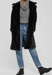 Manteau en fausse fourrure noire à coupe décontractée, grand col, poches, porté sur un pull en laine gris et un jean en denim bleu clair, associé à des bottes noires.