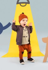 Peuteroutfit bestaande uit een donkerpaarse zip-up jas, een beige shirt met een kattenafbeelding, oranje broek met zwarte vlekken en zwarte schoenen.