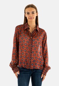 Blusa roja con estampado, de mangas largas, con botones en la parte delantera y cuello puntiagudo. Presenta un diseño geométrico en azul y crema sobre una tela texturada.