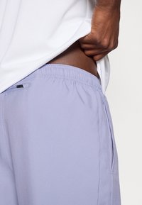 Άτομο που σηκώνει ένα λευκό πουκάμισο, αποκαλύπτοντας σκούρο δέρμα και μέση με ελαστικές κοντές σαλοπέτες σε ανοιχτό μωβ χρώμα που διαθέτουν φερμουάρ στην πλευρά.