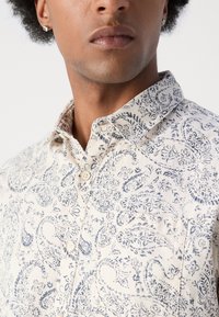 Camisa de botones en tela color crema con un patrón de paisley floral azul, cuello y un único bolsillo en el pecho con un acabado texturizado.