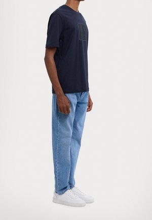 Uomo con maglietta blu scuro con logo discreto, jeans azzurri e sneakers bianche, in piedi davanti a uno sfondo bianco uniforme.