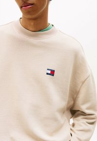 Hellbeige Sweatshirt mit geripptem Kragen und strukturiertem Material. Verfügt über ein kleines, mehrfarbiges Logo-Patch auf der linken Brust.