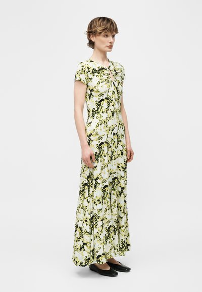 Γυναίκα που στέκεται με μακρύ φόρεμα κίτρινου, μαύρου και λευκού με floral σχέδιο, κοντά μανίκια και ρούχα στο λαιμό, φοράει μαύρες μπαλαρίνες μπροστά σε ένα απλό φόντο.