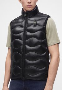 Homme portant un gilet sans manches matelassé noir par-dessus une chemise beige clair à manches courtes, montrant la fermeture éclair frontale du gilet et l’étiquette du logo.
