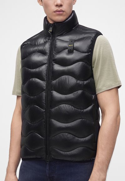 Hombre con chaleco negro acolchado sin mangas sobre una camisa de manga corta beige claro, mostrando la cremallera frontal del chaleco y la etiqueta con el logo.