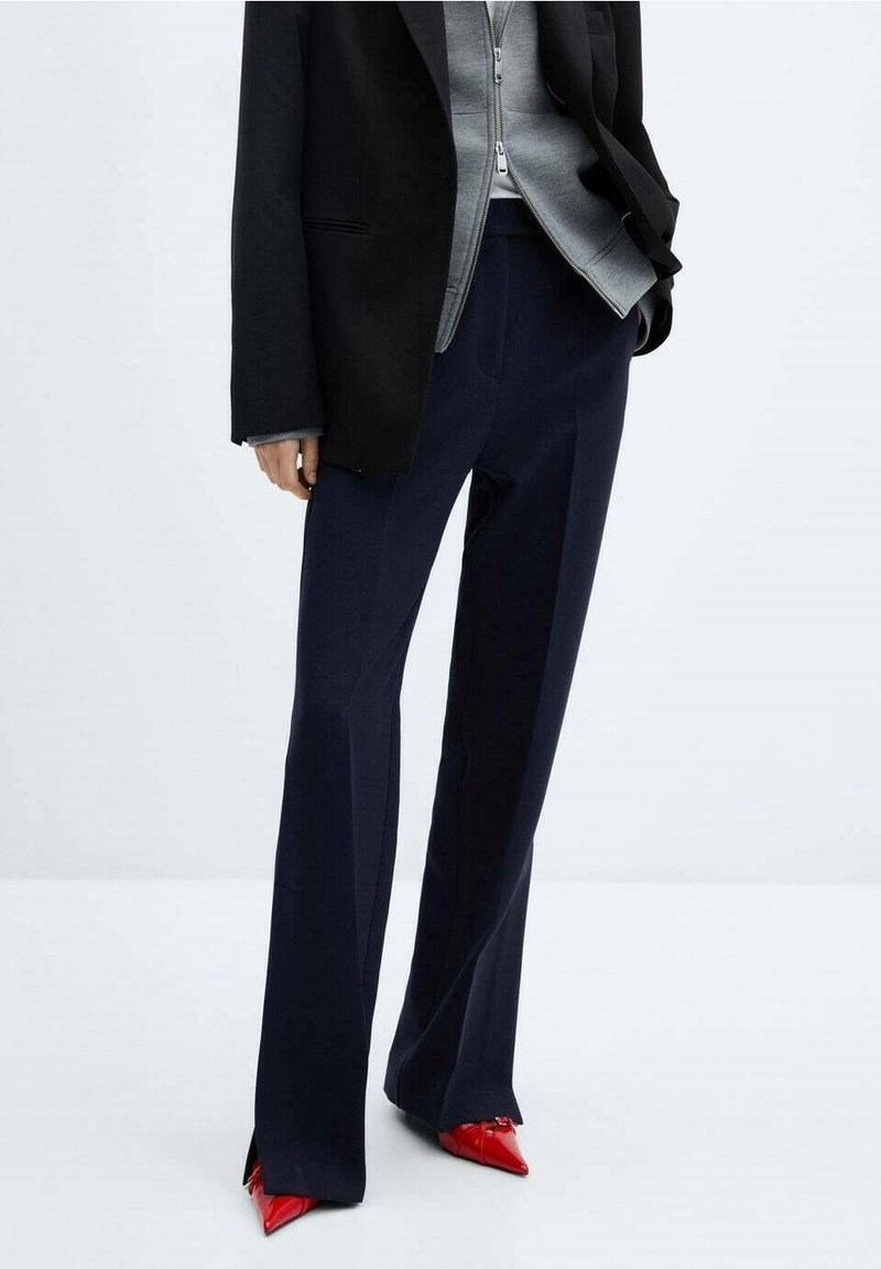 Personne portant un pantalon large bleu marine, des talons hauts rouges pointus, une veste zippée grise et un blazer noir, debout devant un fond uni.