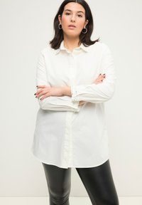 Hvid skjorte med krave, der har dekorative detaljer, lange ærmer med knapdetaljer, parret med sorte læderbukser.