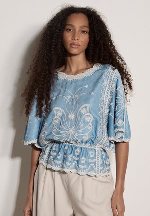 Frau mit langen lockigen Haaren, die eine hellblaue Bluse mit weißer Blumenstickerei und beigen Hosen trägt, steht vor einer schlichten Wand.