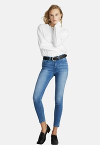 Jeans attillati di un blu chiaro in denim con una texture liscia, abbinati a una camicia bianca a maniche lunghe e una cintura nera. Scarpe nere senza lacci.