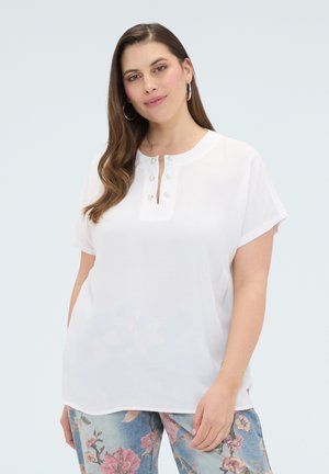 Kvinde med langt brunt hår iført hvid kortærmet bluse med perleknapper og blå bukser med blomsterprint, stående mod en lys baggrund.