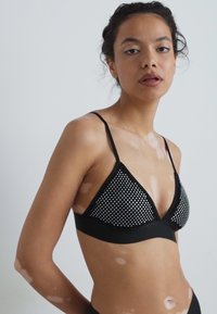 Sujetador triangular negro con cobertura de malla que presenta pequeños remaches plateados, tirantes delgados ajustables y una banda suave debajo del busto.